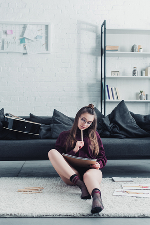 beautiful designer sitting on carpet near couch and holding pencil and notebook at homeの写真素材