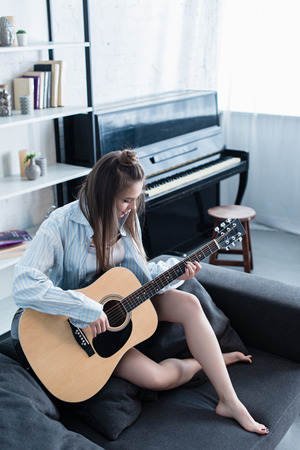 attractive musician sitting on couch and playing acoustic guitar in living roomの写真素材