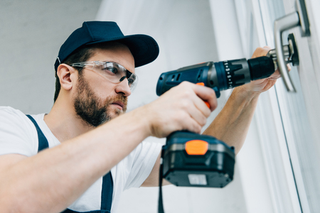 adult repairman in goggles fixing window handle by electric drillの写真素材