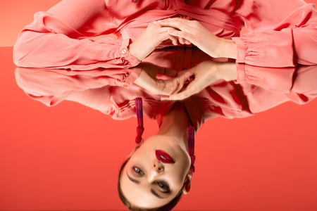 cropped view of beautiful woman in blouse looking at mirror reflection isolated on living coral