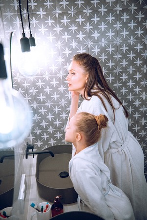 beautiful woman standing with daughter in white bath robes in bathroomの写真素材