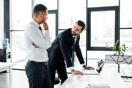 confident businessman standing near desk and talking with coworker in modern officeの写真素材