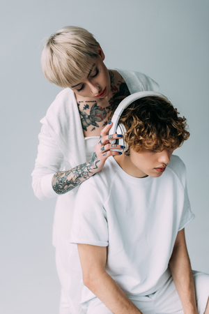 blonde tattooed girl holding headphones near man with curly hair listening music isolated on greyの写真素材