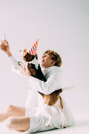 happy man with curly hair holding cupcake above head near beagle dog in party cap on grey backgroundの写真素材