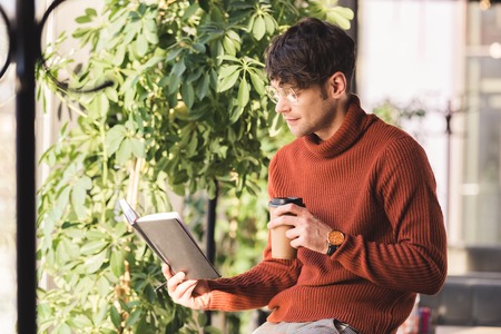 thoughtful man in glasses looking at notebook and holding disposable cup in cafe