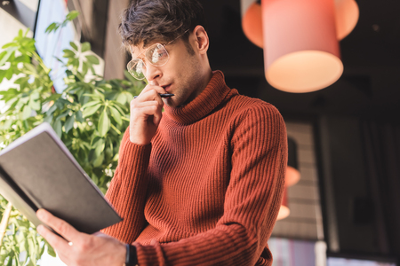 pensive man in glasses holding notebook while standing in cafeの写真素材