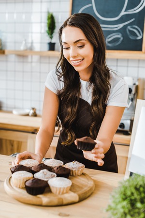 selective focus of attractive waitress standing behind bar counter and putting cupcakes on wooden tray in coffee houseの写真素材