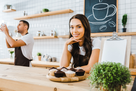 attractive brunette waitress standing behind bar with cupcakes on wooden tray wile barista holding cup in coffee houseの写真素材