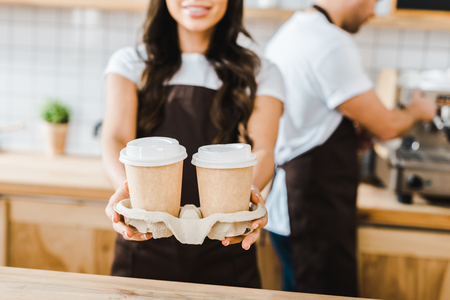 cropped view of brunette cashier standing and holding paper cups wile barista  working in coffee houseの写真素材