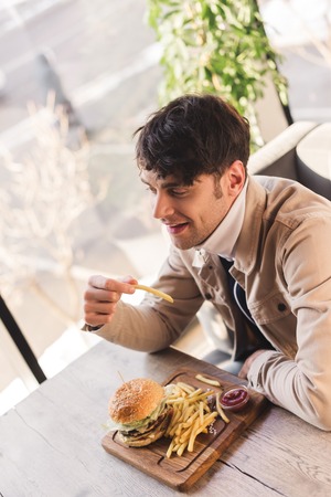 cheerful man holding french fry near tasty burger on cutting board in cafeの写真素材