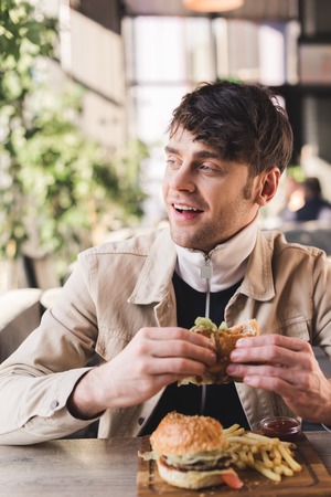 selective focus of smiling young man holding tasty hamburger near french fries on cutting board in...