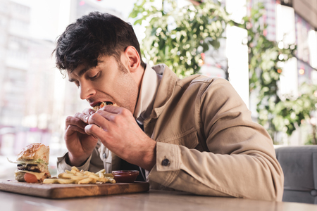 selective focus of young man eating tasty hamburger near french fries on cutting board in cafeの写真素材