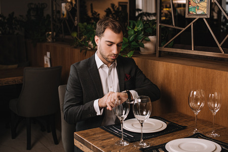 handsome man in suit waiting for girlfriend in restaurant and looking at watchの写真素材
