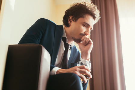 adult sad businessman in formal wear holding glass of whiskey in hotel roomの写真素材