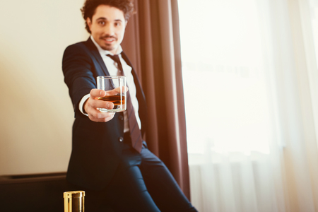 selective focus of smiling businessman in formal wear holding glass of whiskey in hotel roomの写真素材