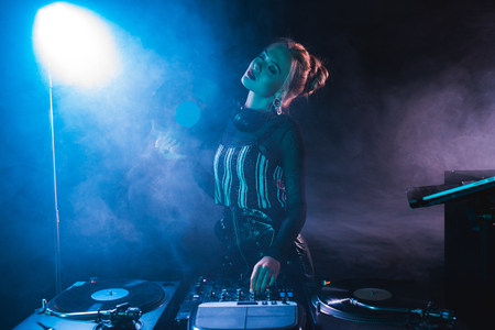 blonde dj woman standing with closed eyes near dj equipment and holding retro vinyl record in nightclub with smokeの写真素材