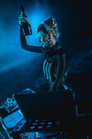 cheerful blonde dj woman in headphones holding  bottle and smiling near dj equipment in nightclub with smokeの写真素材