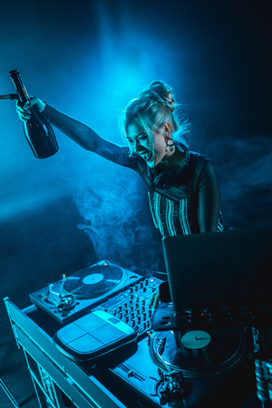 cheerful blonde dj woman holding  bottle and smiling near dj equipment in nightclub with smokeの写真素材