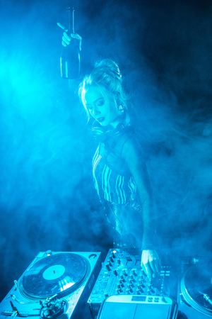 blonde dj woman in headphones holding  bottle near dj equipment in nightclub with smokeの写真素材