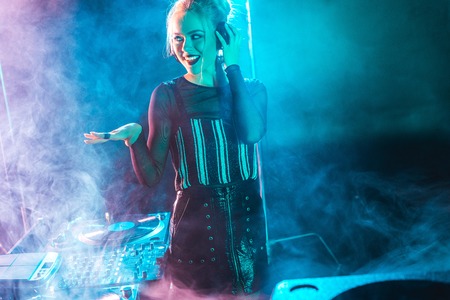 happy dj woman with blonde hair listening music and touching headphones in nightclub with smoke