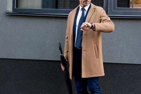 cropped view of businessman looking at watch and holding umbrellaの写真素材