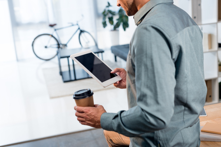 cropped view of businessman holding disposable cup and digital tablet with blank screen in officeの写真素材