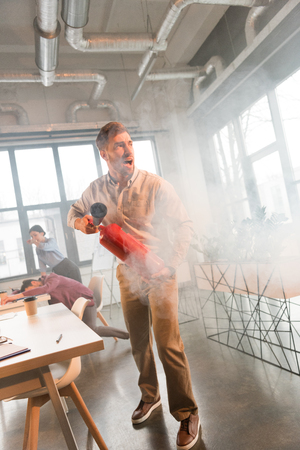 handsome businessman holding extinguisher and screaming in office with smoke near colleaguesの写真素材