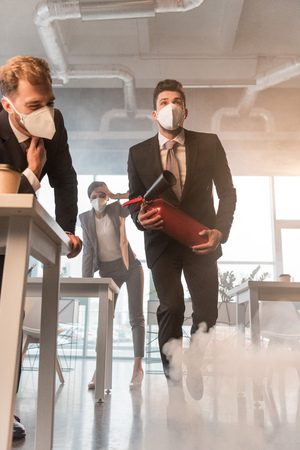 businessman in mask holding extinguisher near coworkers in office with smokeの写真素材