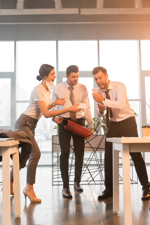 terrified businessman holding extinguisher near yelling coworkers looking at smartphone in officeの写真素材