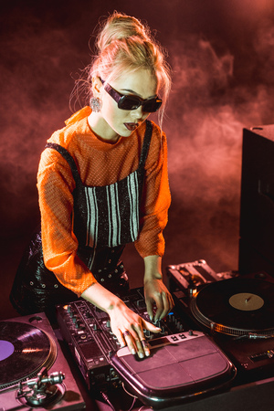 blonde dj girl in sunglasses touching dj equipment in nightclub with smokeの写真素材