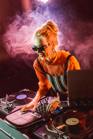 cheerful dj girl in sunglasses touching dj equipment in nightclub with smokeの写真素材