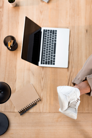 top view of laptop with blank screen with male legs in white sneakers on tableの写真素材