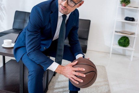 cropped view of businessman in glasses and suit holding basketball in hands while sitting on coffee tableの写真素材
