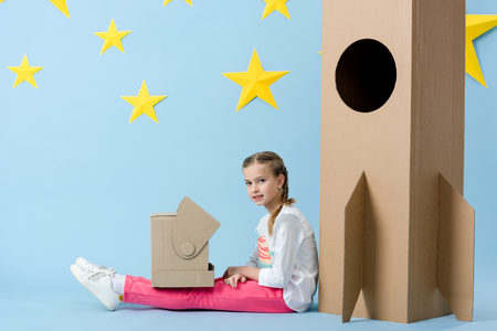 Interested kid with helmet sitting near cardboard rocket and looking at camera on blue starry backgroundの写真素材