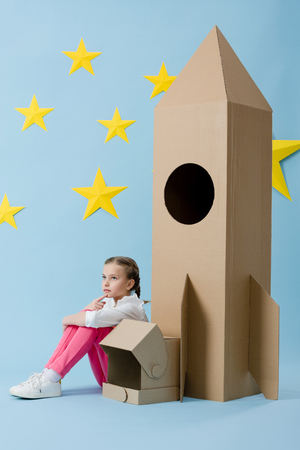 Dreamy kid with helmet sitting near cardboard rocket on blue starry backgroundの写真素材