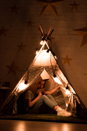 Preteen kid in cardboard helmet sitting in wigwam in dark roomの写真素材