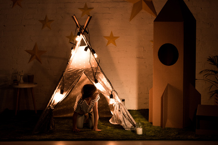 Kid standing on all fours in wigwam and looking at cardboard rocket in dark roomの写真素材