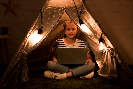 Happy kid using laptop while sitting in wigwam in dark roomの写真素材