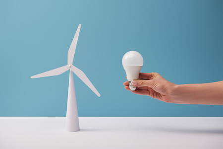cropped view of woman holding light bulb over white table with windmill model on blue background