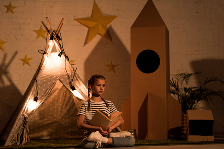 Relaxed kid sitting in lotus pose and reading book in dark room