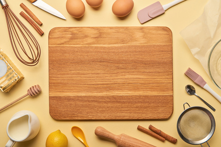 top view of empty wooden chopping board with cooking utensils and ingredients on yellow background