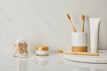 toothpaste in tube, holder, bamboo toothbrushes, cosmetic cream and ear sticks in glass on white marble backgroundの写真素材