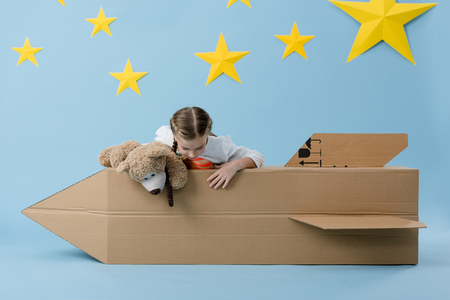 Kid with teddy bear playing with cardboard rocket on blue background with starsの写真素材