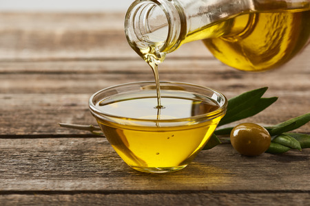pouring oil from bottle into glass bowl, olive tree leaves and olive on wooden surfaceの写真素材