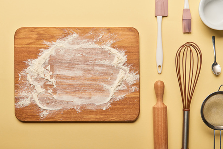 top view of wooden chopping board with flour and cooking utensils on yellow background