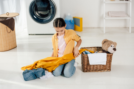 child in yellow shirt and jeans sitting near basket and holding clothes in laundry roomの写真素材