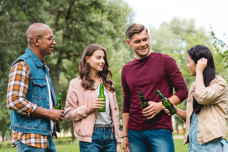 happy multicultural friends holding bottles with beer while standing in park
