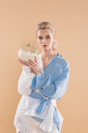 beautiful woman holding fish bowl with sand and small green plant while standing in eco clothing isolated on beige, environmental saving conceptの写真素材