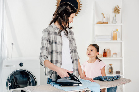 mother ironing and looking to daughter standing near in laundry roomの写真素材
