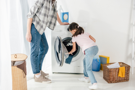 daughter in pink t-shirt putting clothes in washer wile mother standing in laundry roomの写真素材